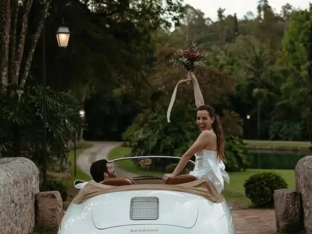 Noivos posam junto a um conversível vintage, ela erguendo o buquê ao vento