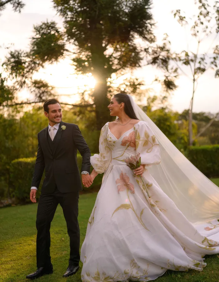 Casal caminha de mãos dadas pelo jardim da Fazenda Fortaleza ao entardecer, vestido com flores pintadas à mão e véu longo ao vento